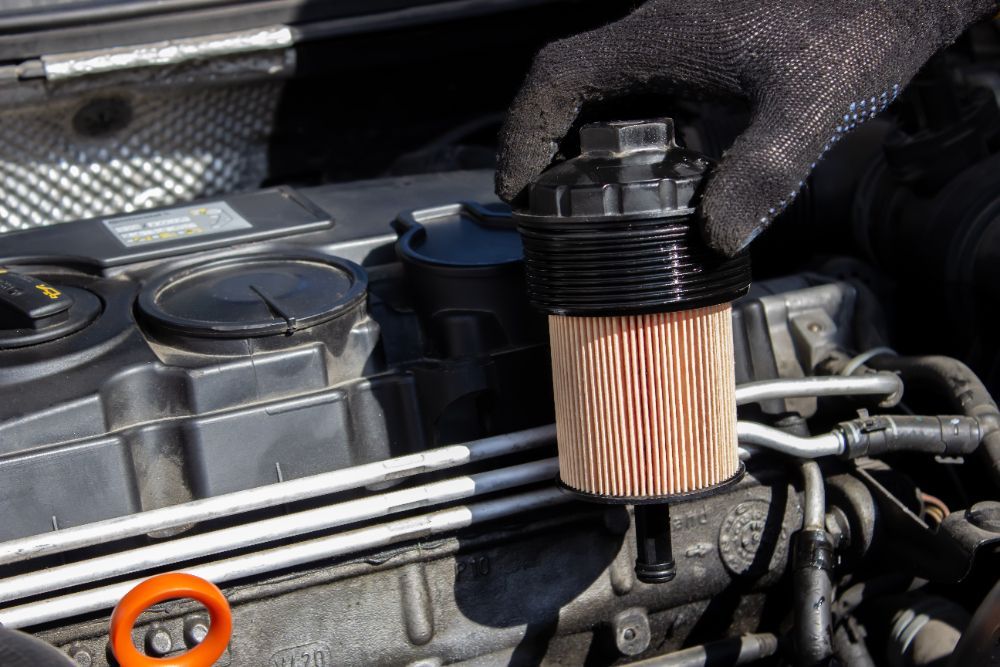 Gloved hand removing a car oil filter from an engine. Black filter cap, tan filter element, engine details.