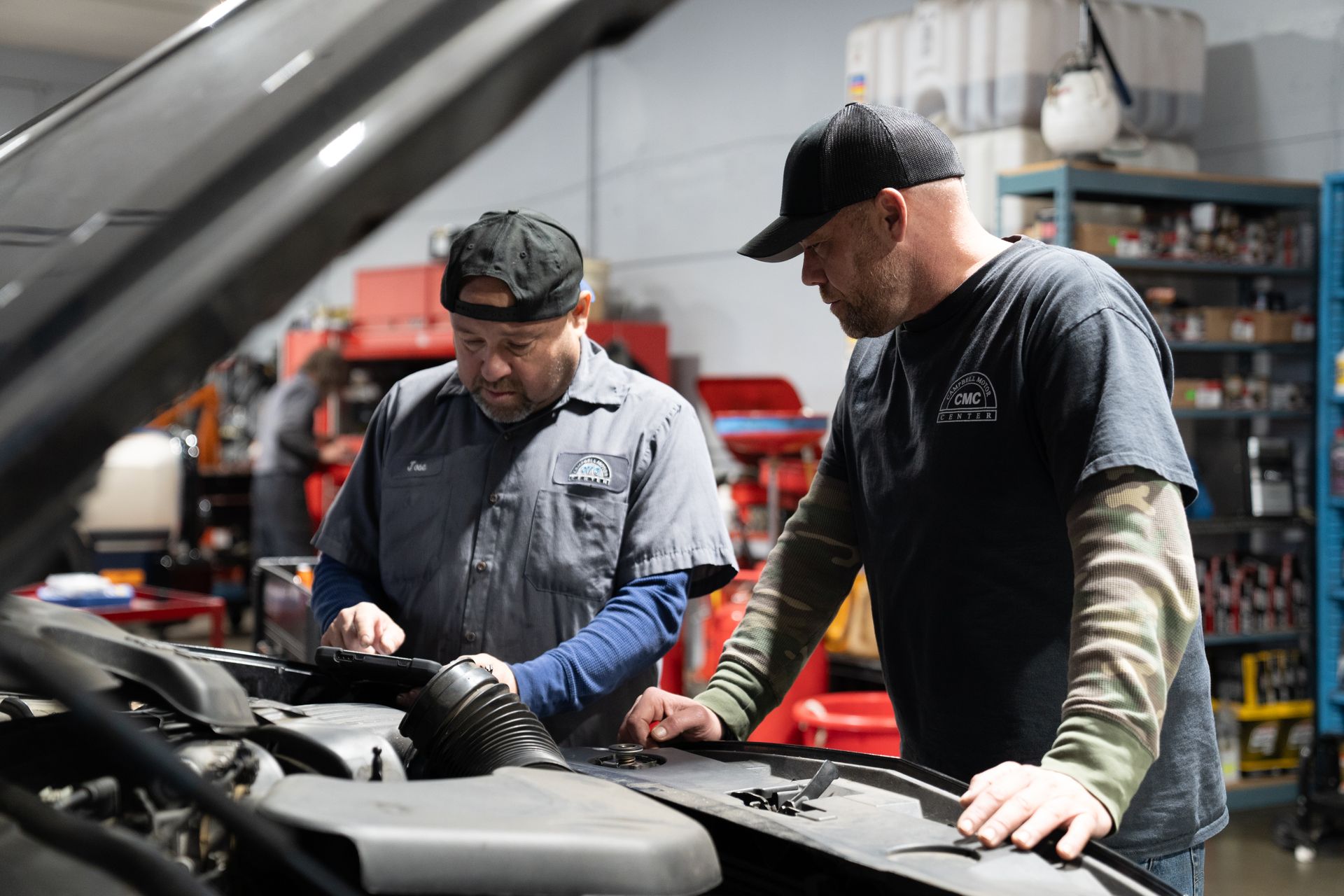 Two men in a garage working on a car engine. One holds a tablet, both wearing hats.