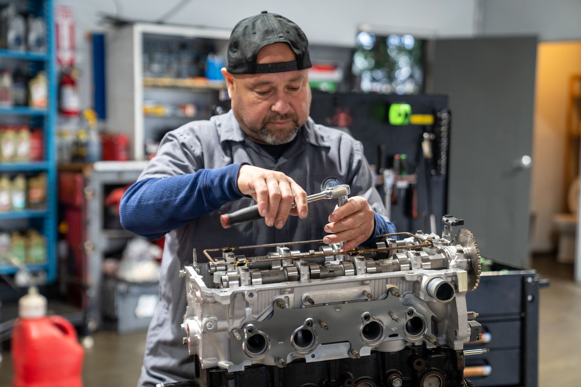 Mechanic in a gray shirt and black cap working on an engine in a garage.