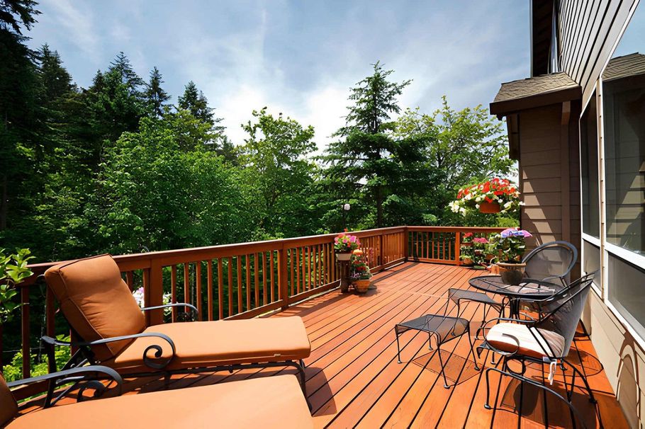 Sunlit wooden deck with orange lounge chairs, patio table, and lush green trees beyond the railing