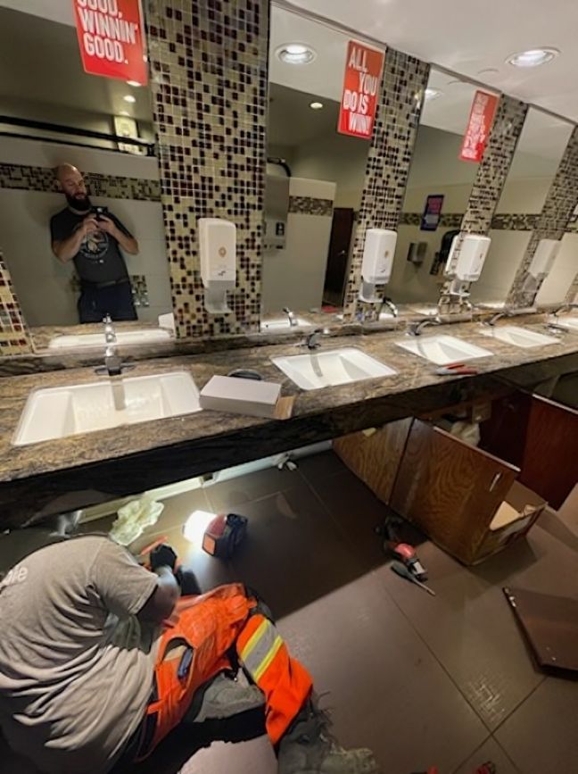 Public restroom with broken sink and mirror; worker in orange pants crouches under counter, inspecting damage.