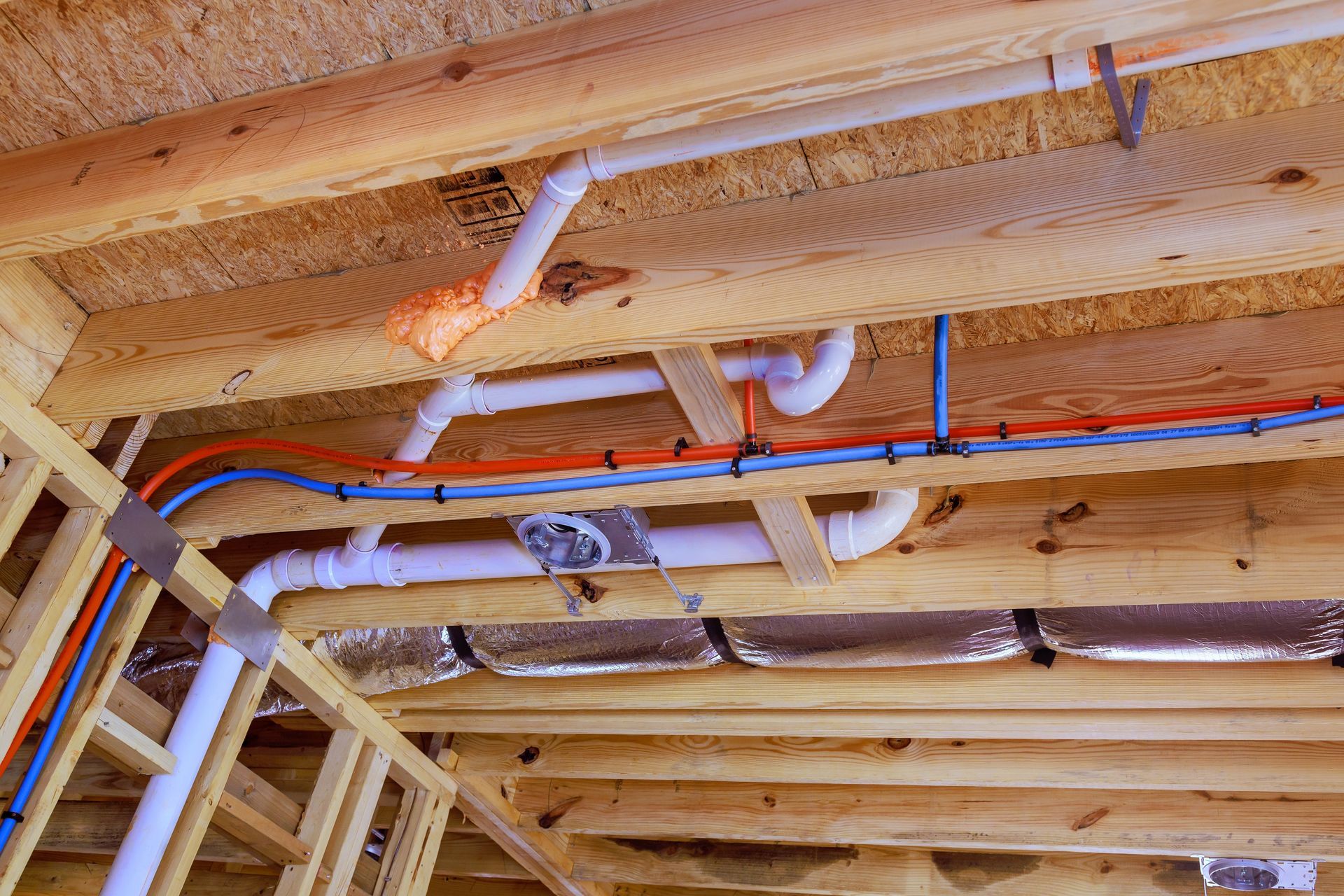 Exposed ceiling framing with white PVC plumbing pipes and red-blue electrical wiring in a house under construction