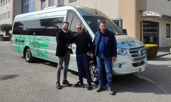 Tre uomini posano accanto a un furgone passeggeri di colore argento e verde in un parcheggio.