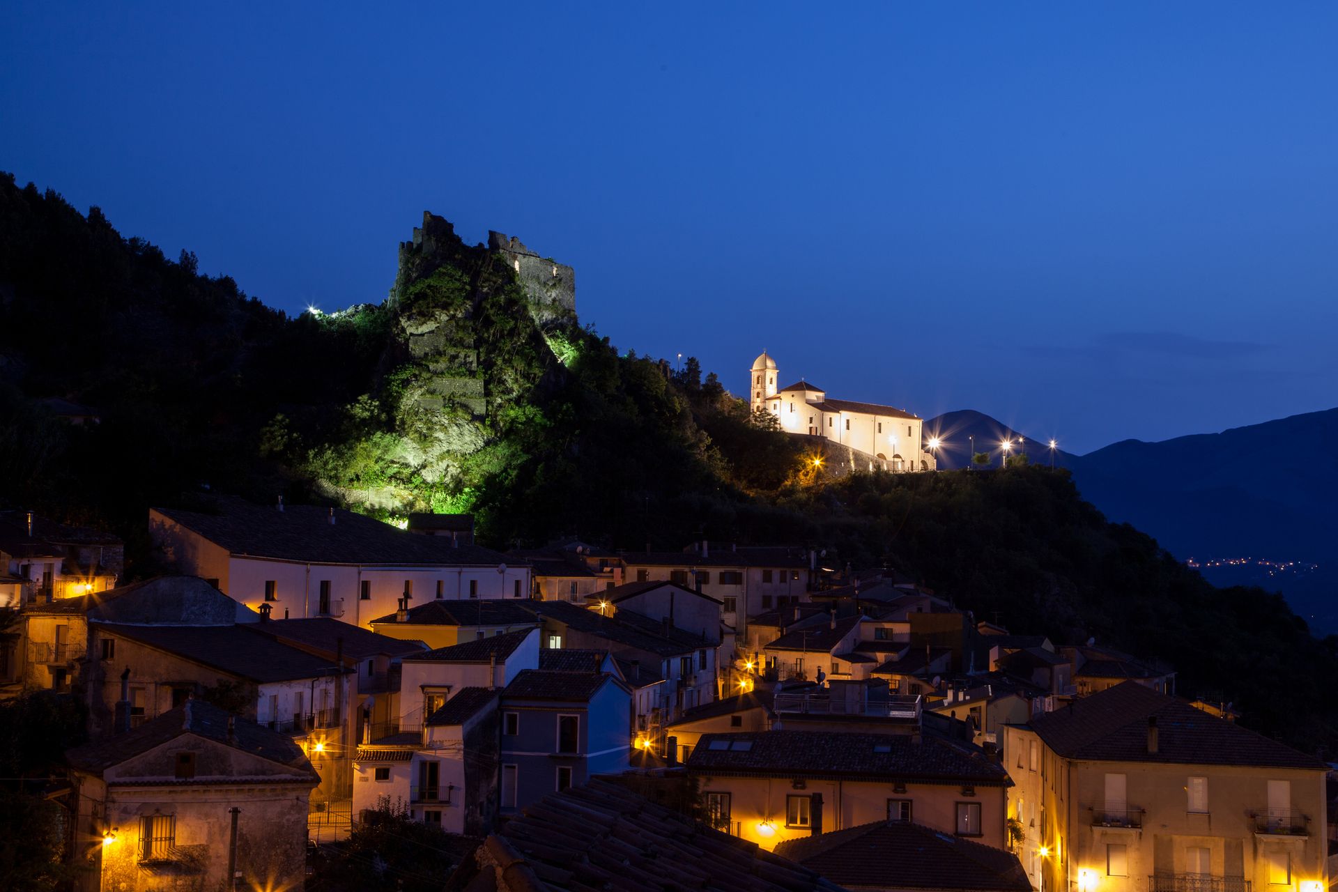 Veduta notturna di un villaggio collinare con luci calde e un castello illuminato in cima alla collina