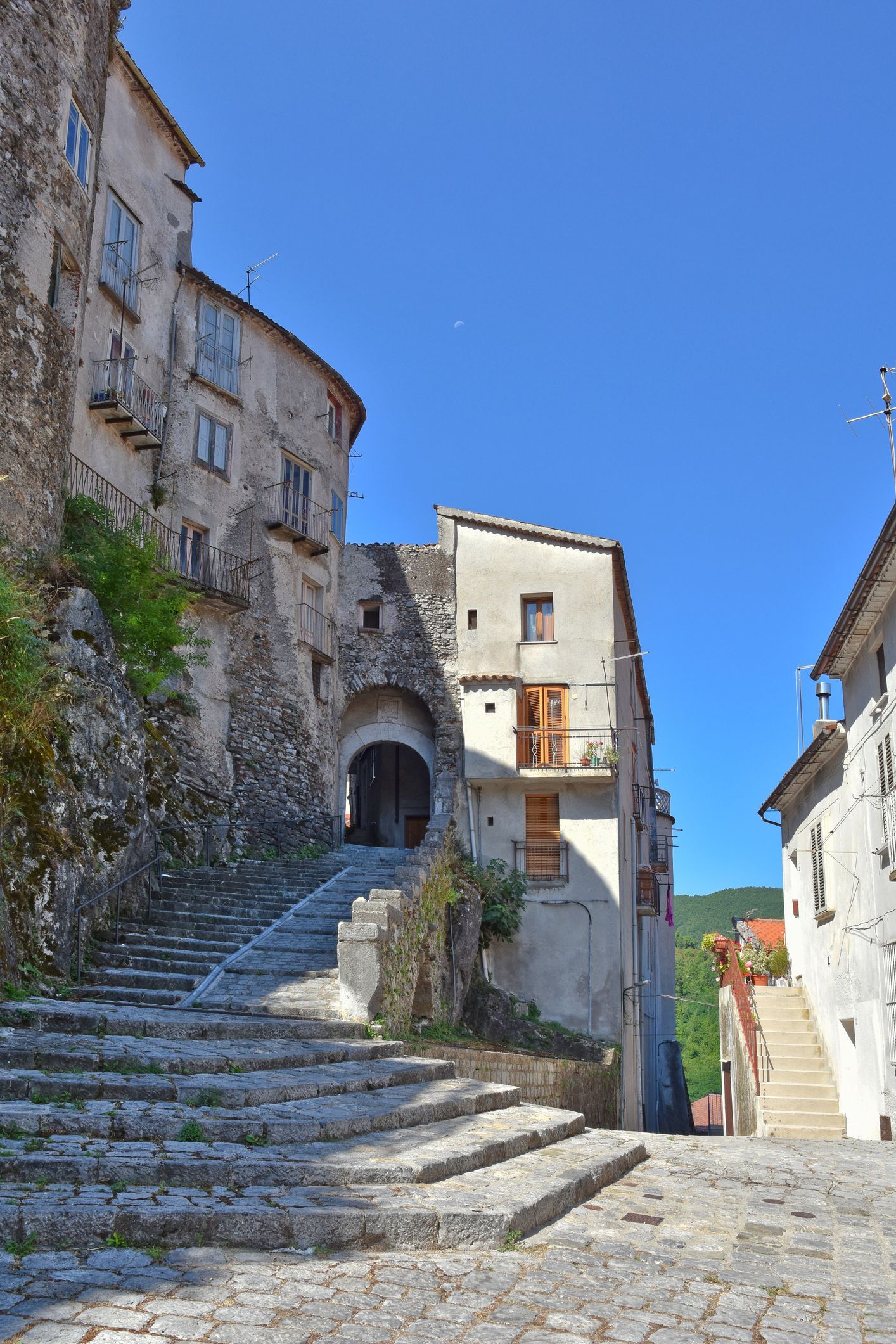 In una stretta via di paese, sotto un cielo azzurro e limpido, una scalinata di pietra si inerpica tra antichi edifici in pietra.