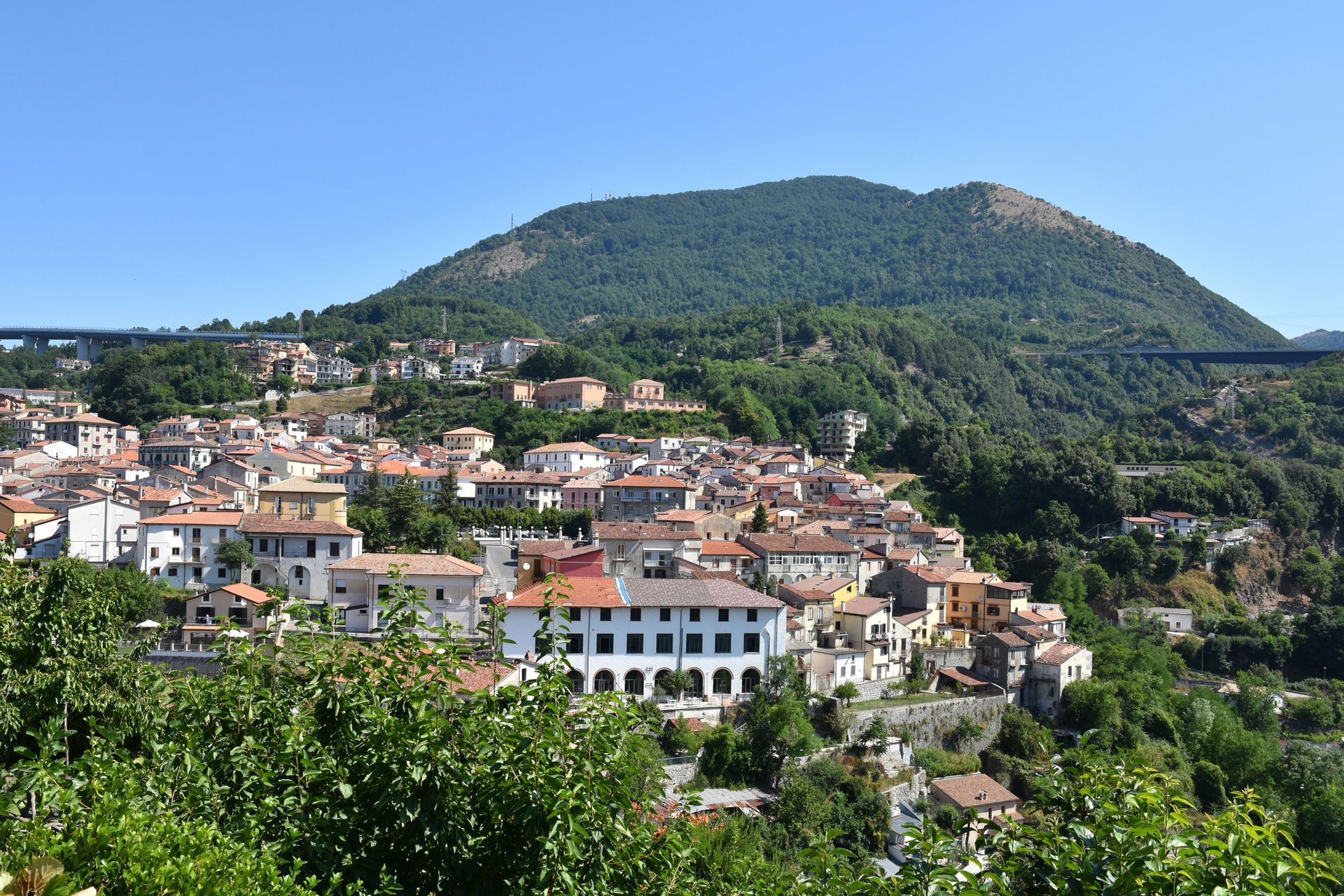 Cittadina dai tetti rossi ai piedi di una verde montagna sotto un cielo azzurro limpido.