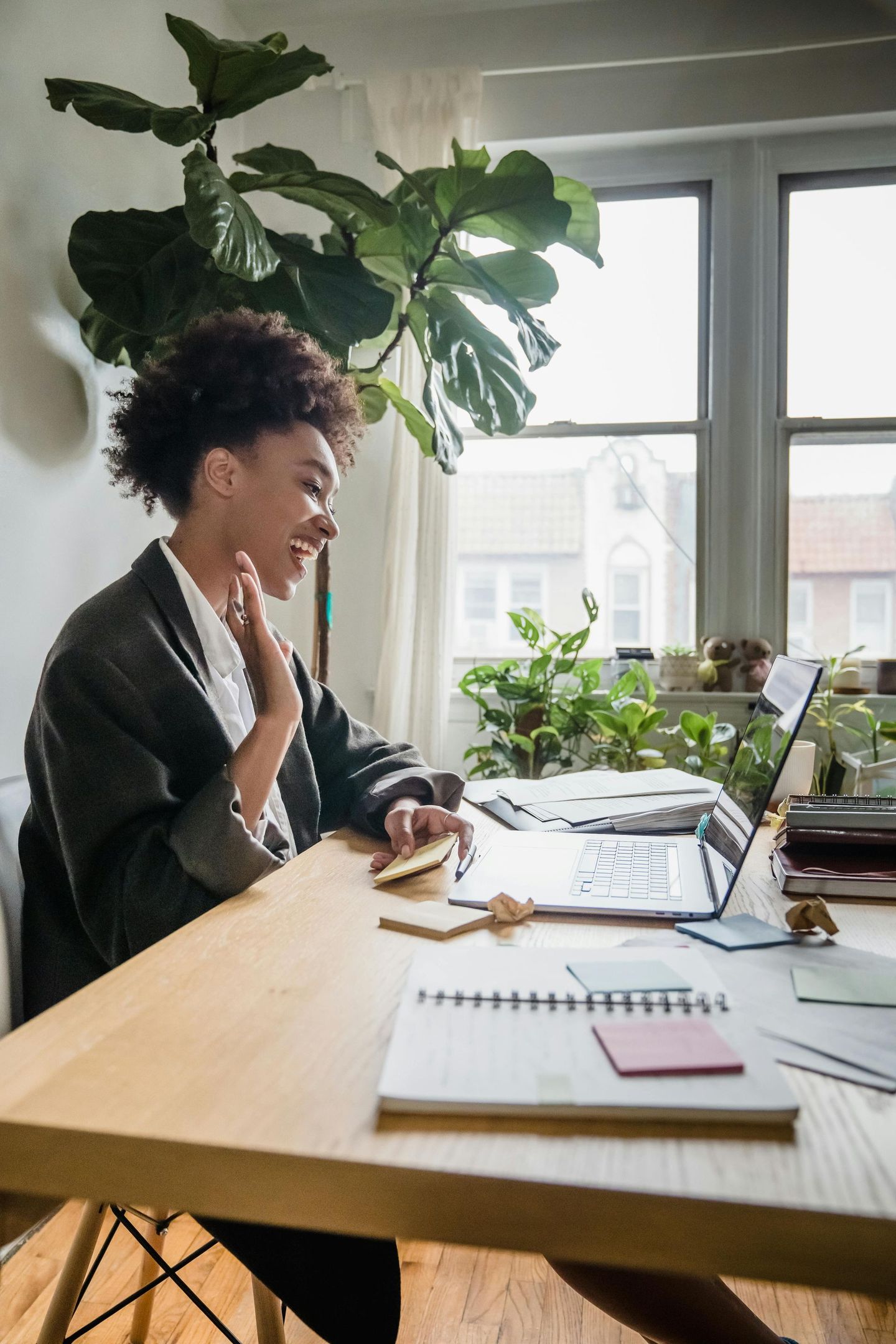Woman waves at laptop during video call, sitting at a wooden desk in a well-lit room.