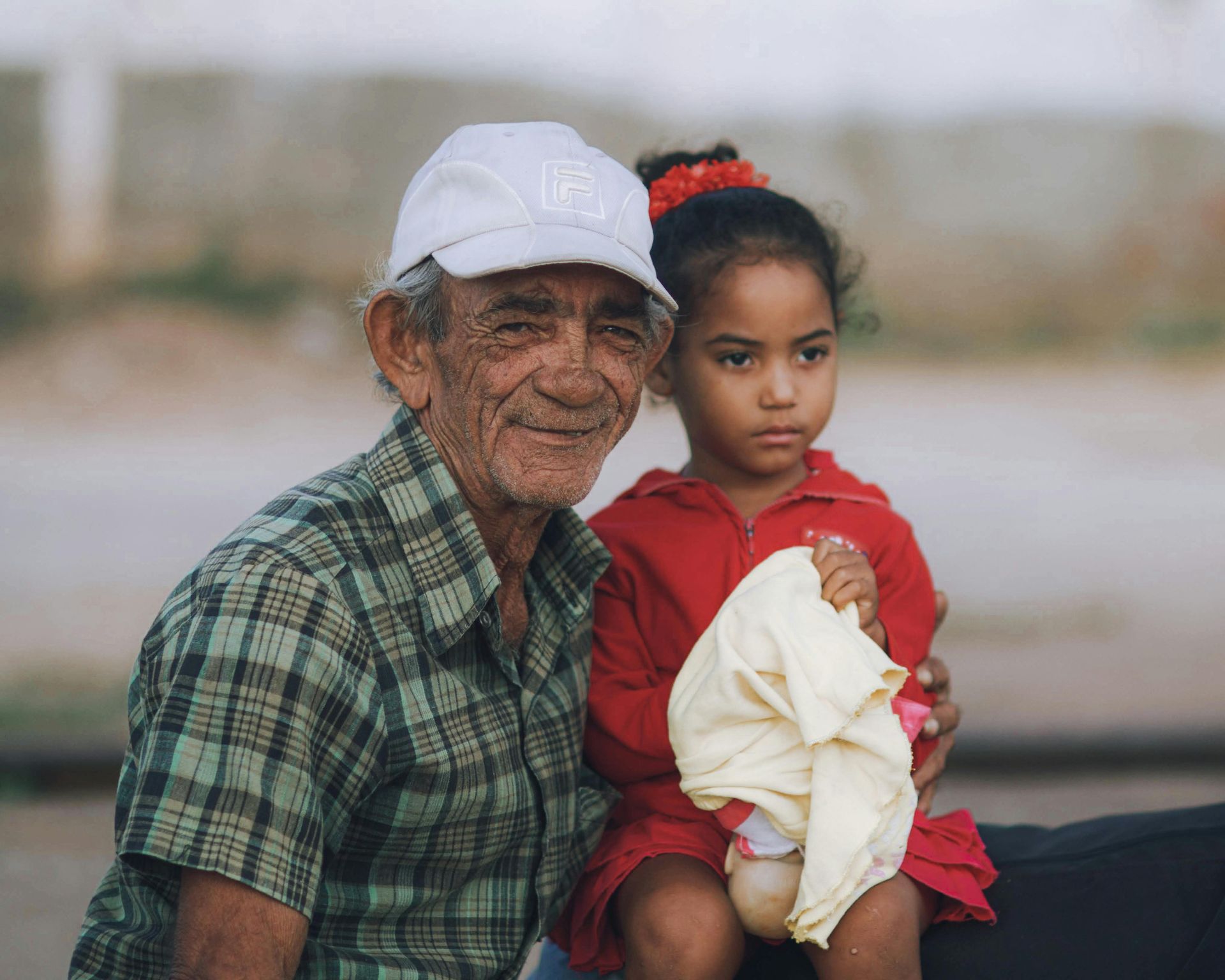 An older man in a plaid shirt and white cap holds a young child in a red shirt, who is holding a yellow cloth.