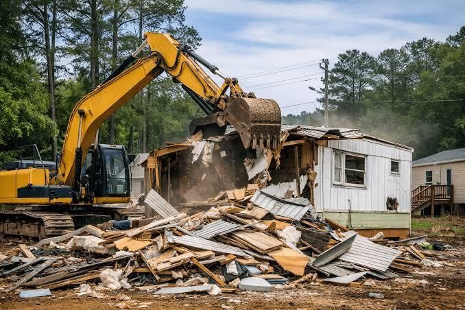 Yellow excavator demolishing a small house, rubble piled in front, wooded neighborhood background