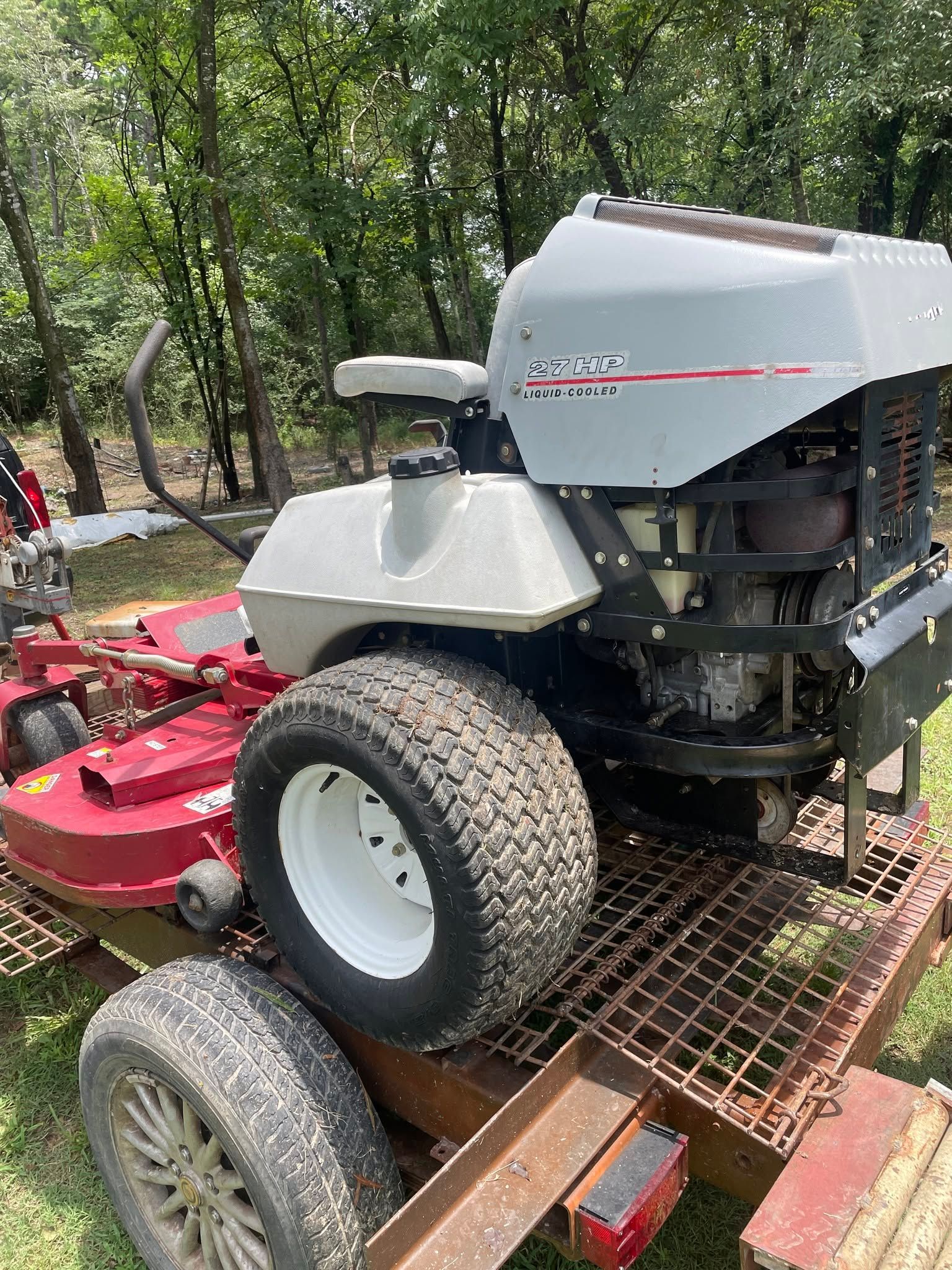 Gray riding lawn mower strapped on a trailer in a wooded area