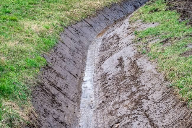 Mud-filled ditch running through grassy ground with sloped, eroded sides