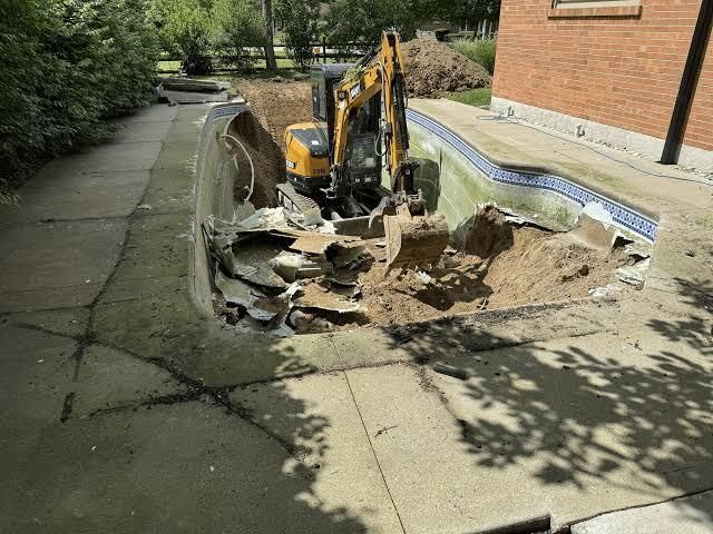 Mini excavator digging up concrete beside a brick building on a tree-lined sidewalk
