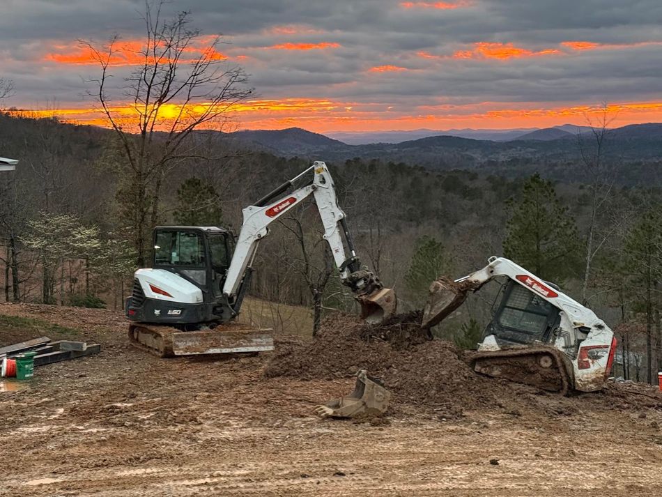 Two excavators working on a dirt site at sunset with hills in the background