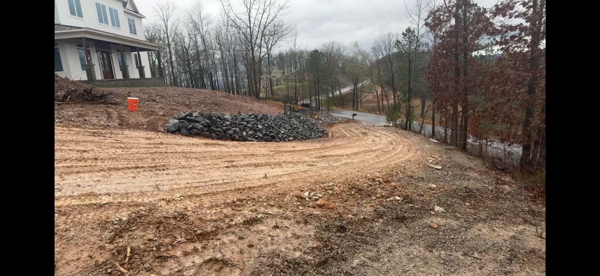 Gravel driveway and bare yard beside a house, with a wood pile and winter trees