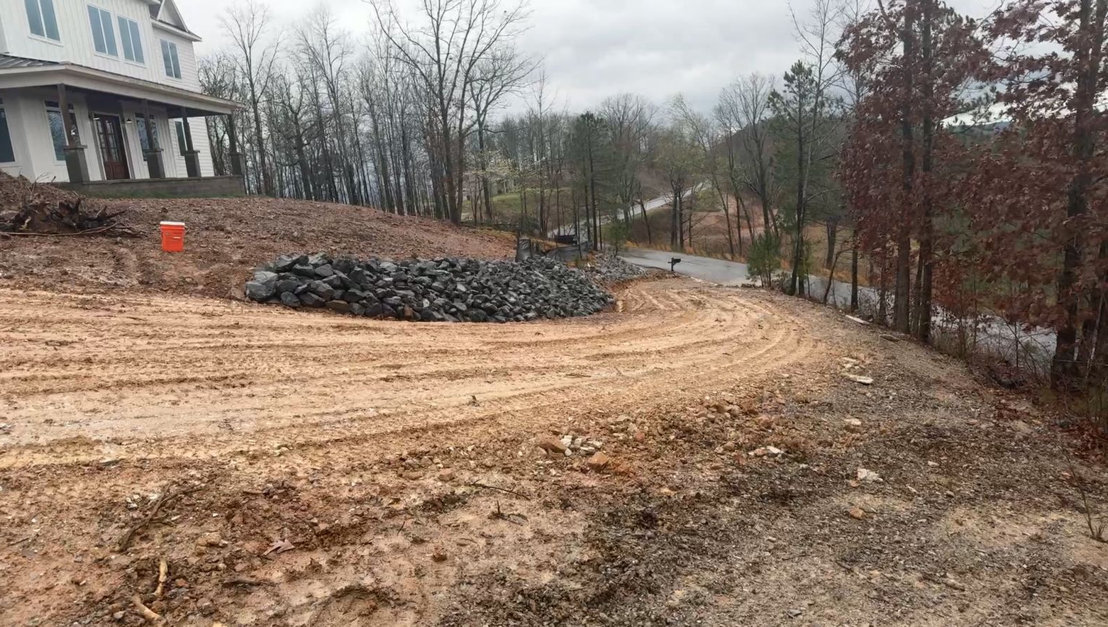 Mud driveway beside a white house, with stacked rocks and leafless trees on a wet, overcast hillside.
