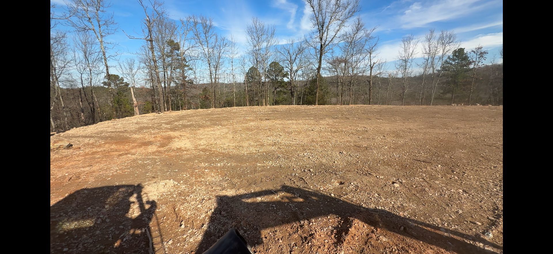 Open dirt clearing bordered by trees under a blue sky, with wooden railing in the foreground.