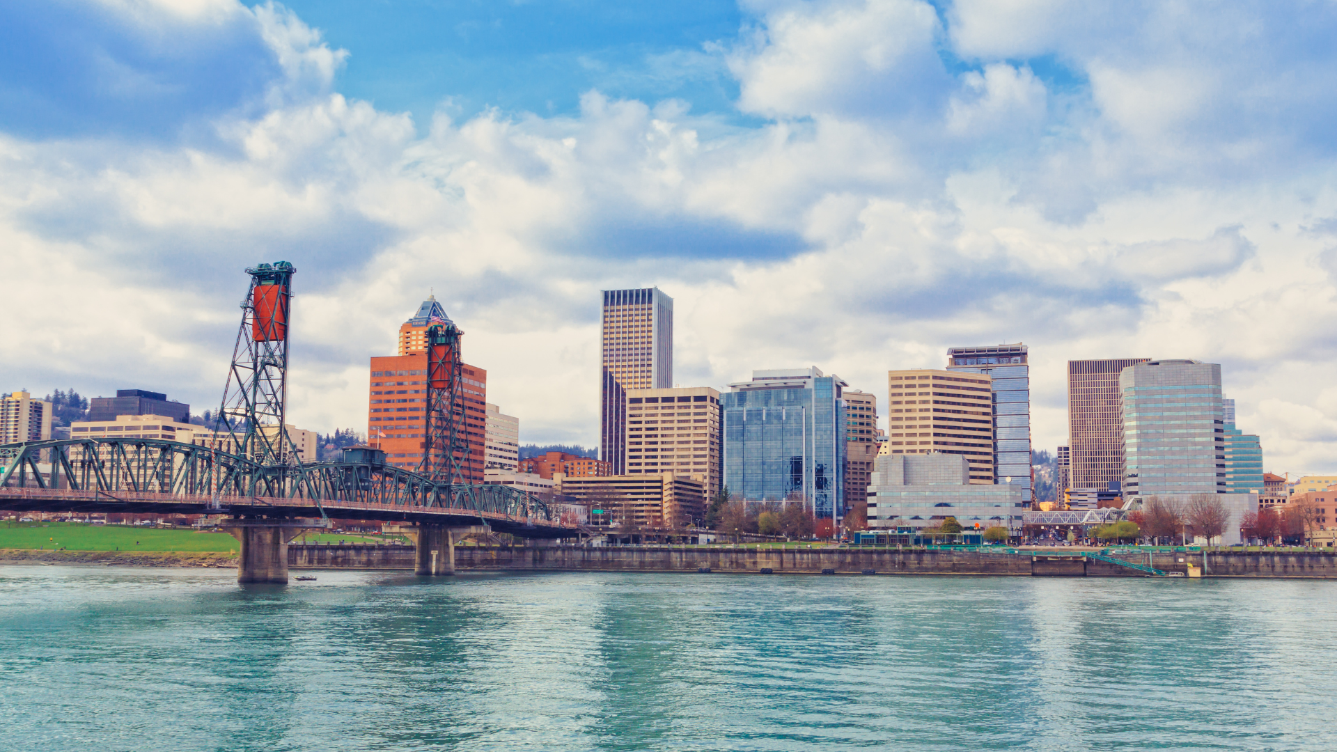 Portland, Oregon skyline with Willamette River and bridge under a cloudy blue sky.