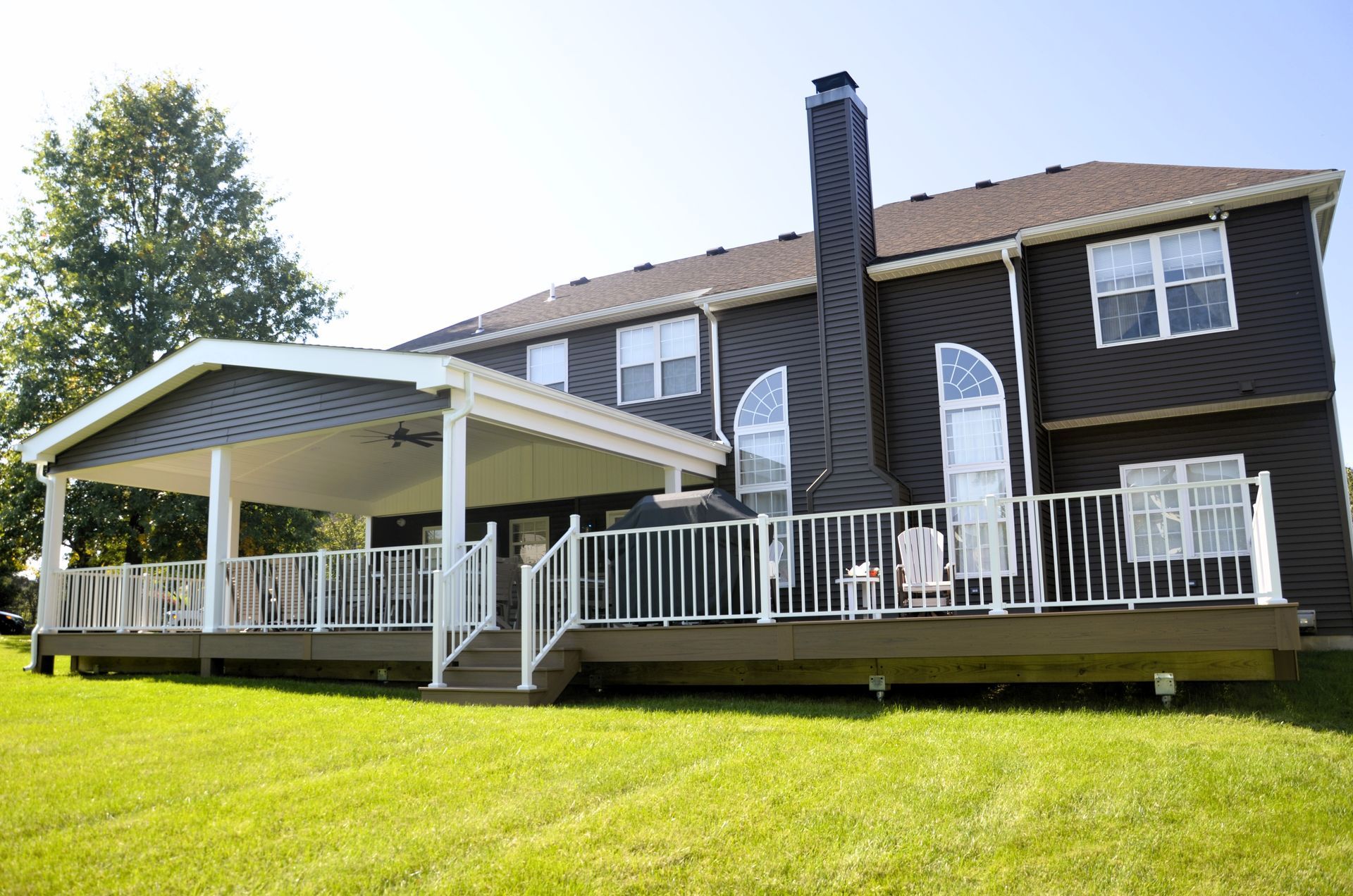 Two-story house with a covered deck, white railing, and a tall chimney on a grassy lawn