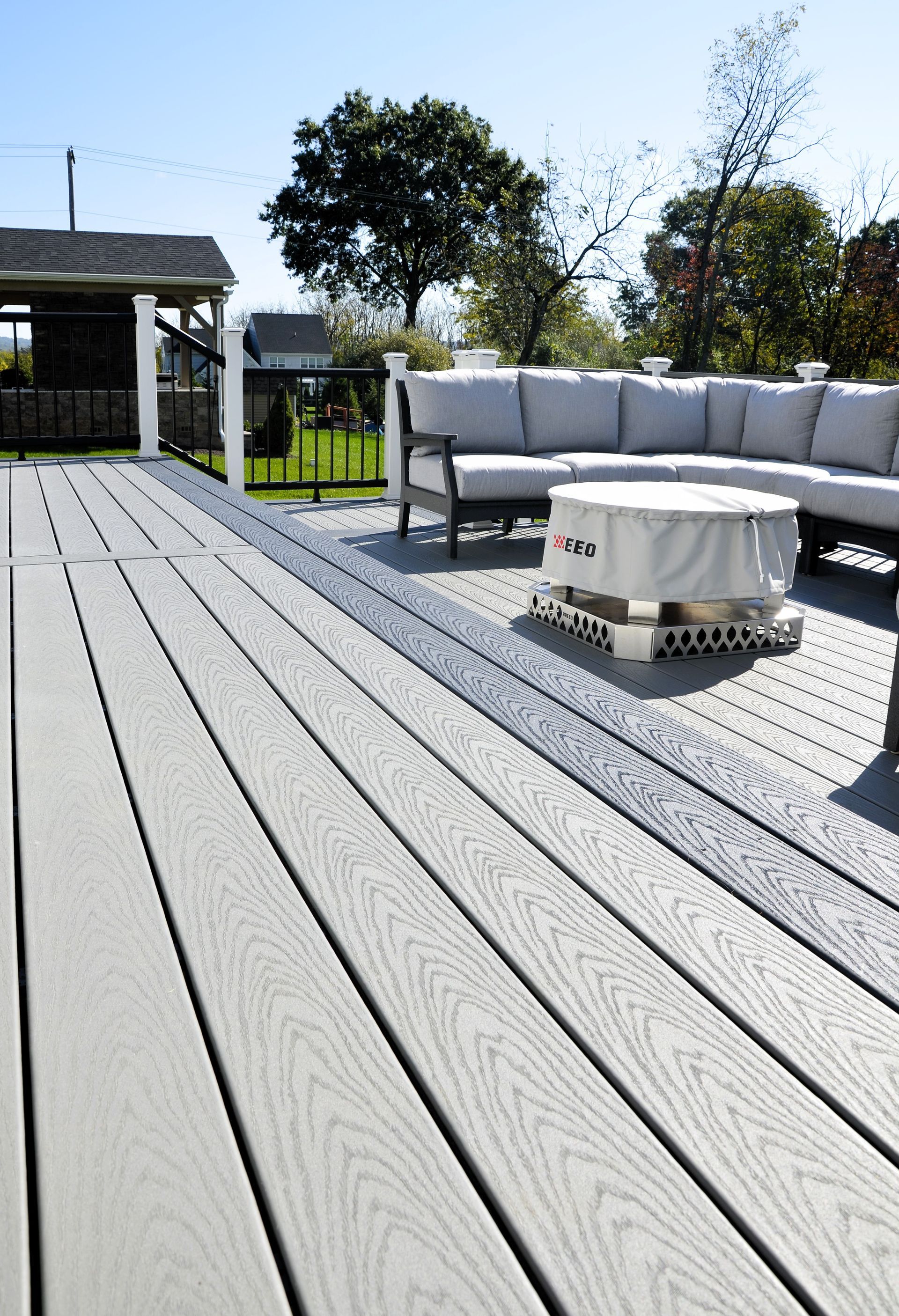 Outdoor deck with gray flooring, white sectional sofa, and fire pit in a sunny backyard