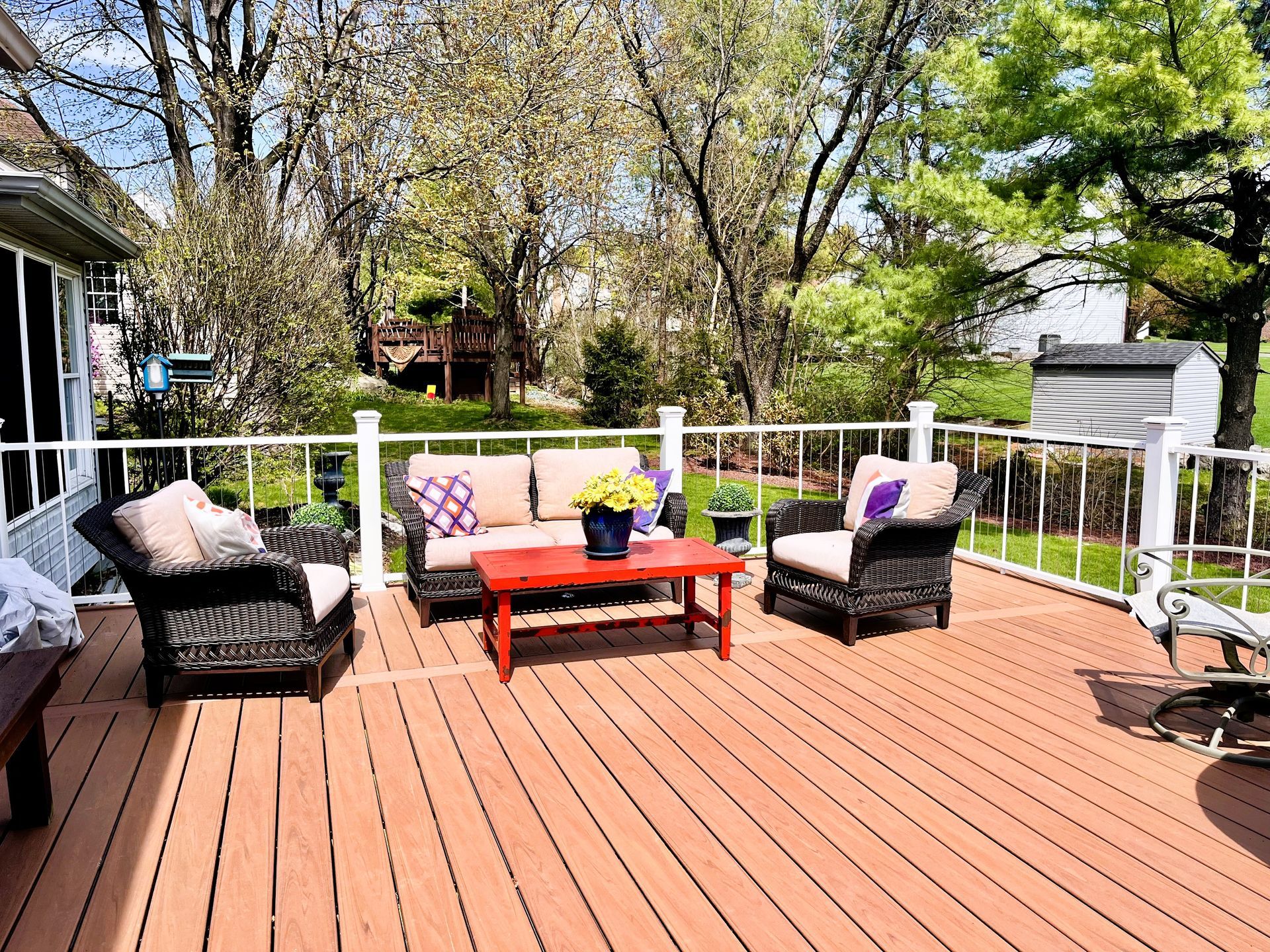 Patio deck with wicker seating, red coffee table, and potted flowers in a sunny backyard garden