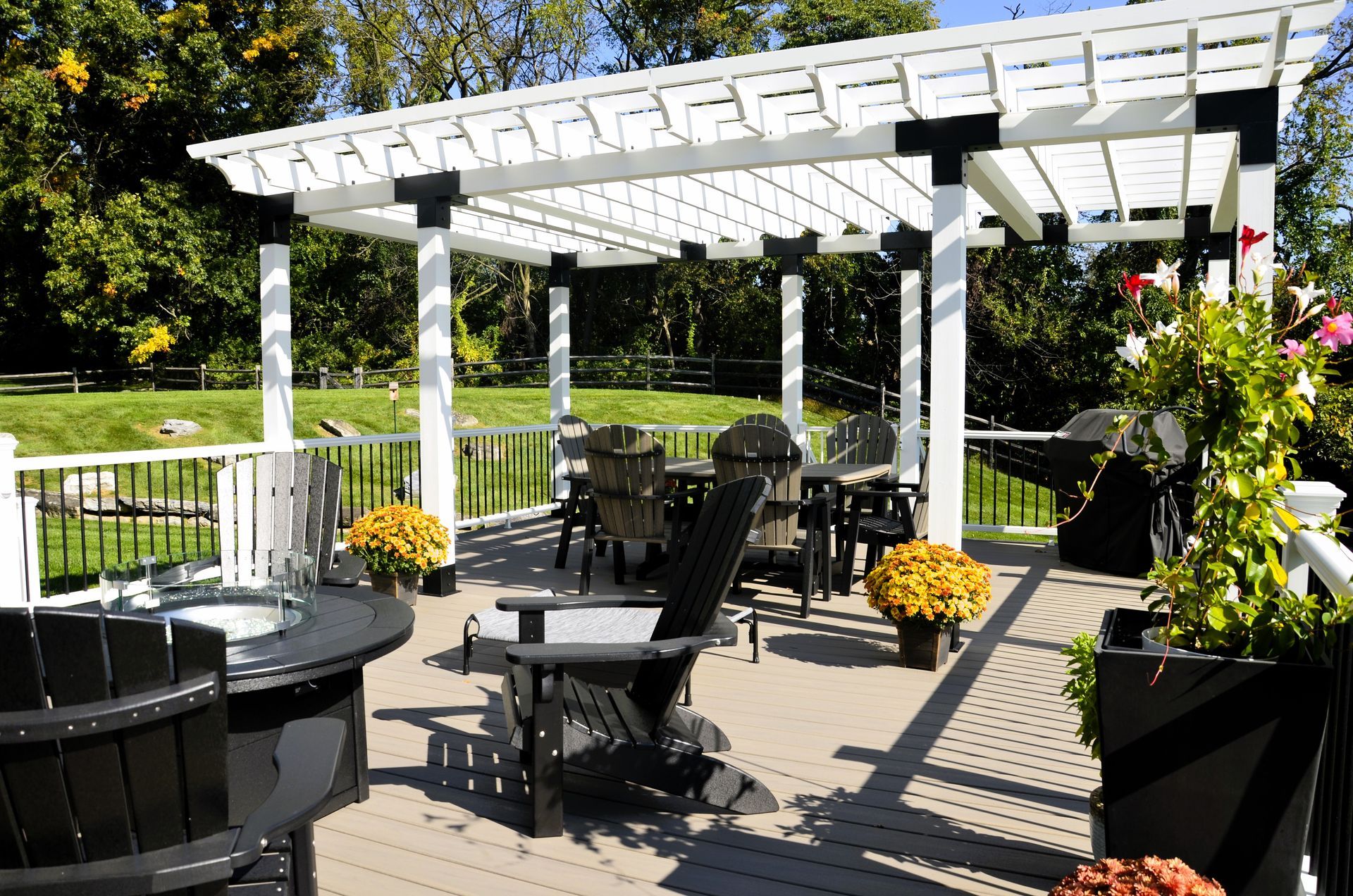 Backyard patio with white pergola, black chairs, round table, and potted flowers in sunlight