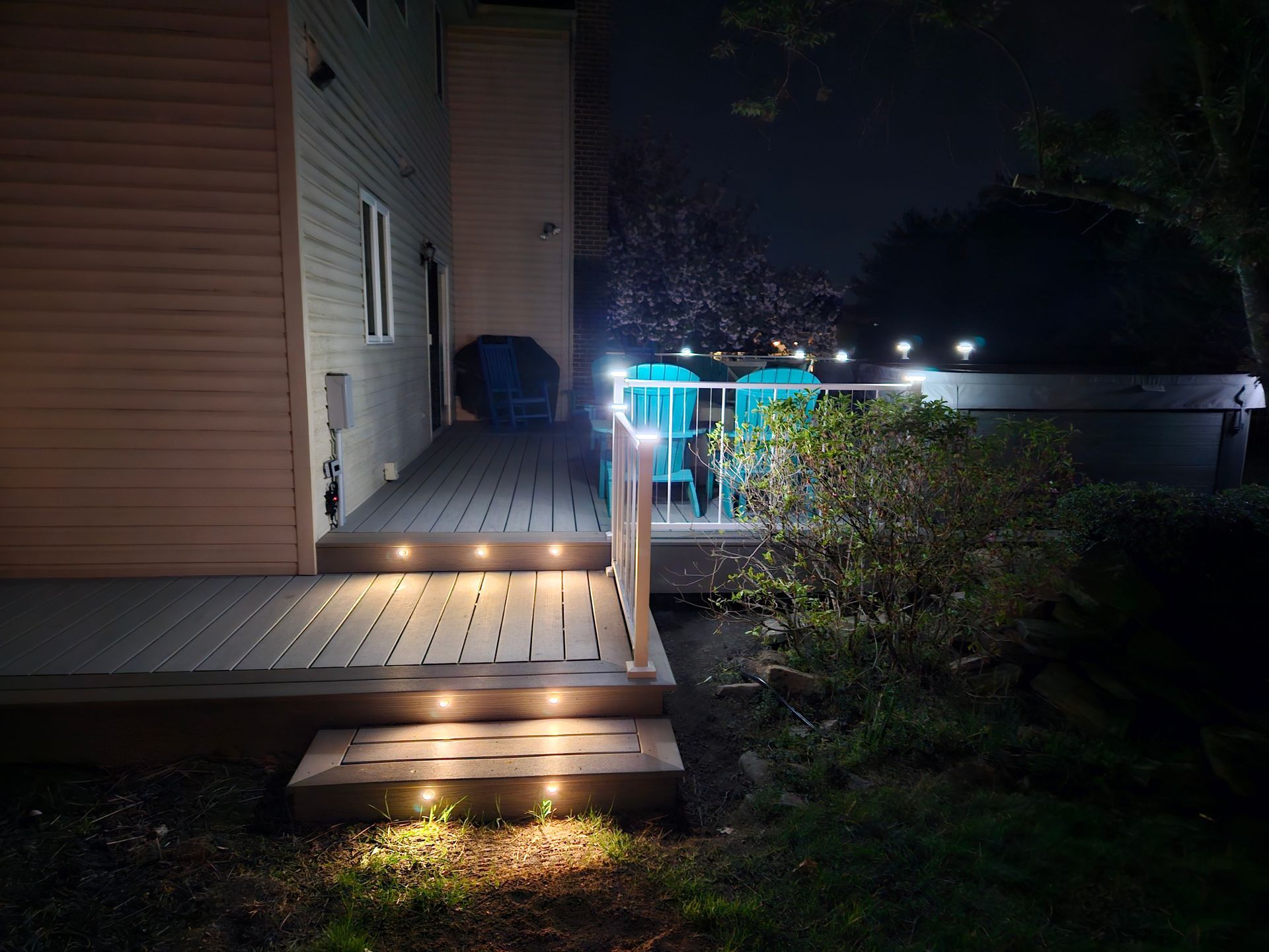 Nighttime exterior of a house with a lit wooden deck, steps, and a glowing blue pool area beyond the hedge