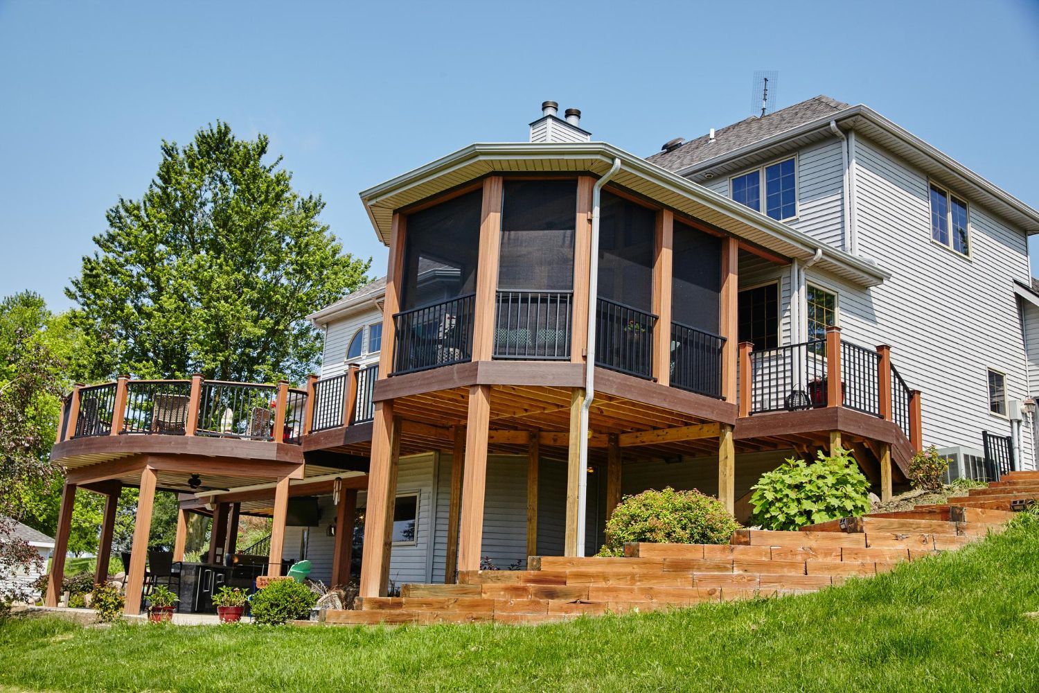 Large modern house with wraparound deck, wood posts, and green lawn on a sunny day