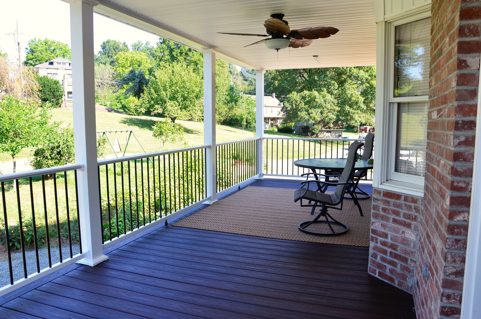 Covered porch with dark wood floor, black railing, rocking chairs, and a ceiling fan overlooking a green yard