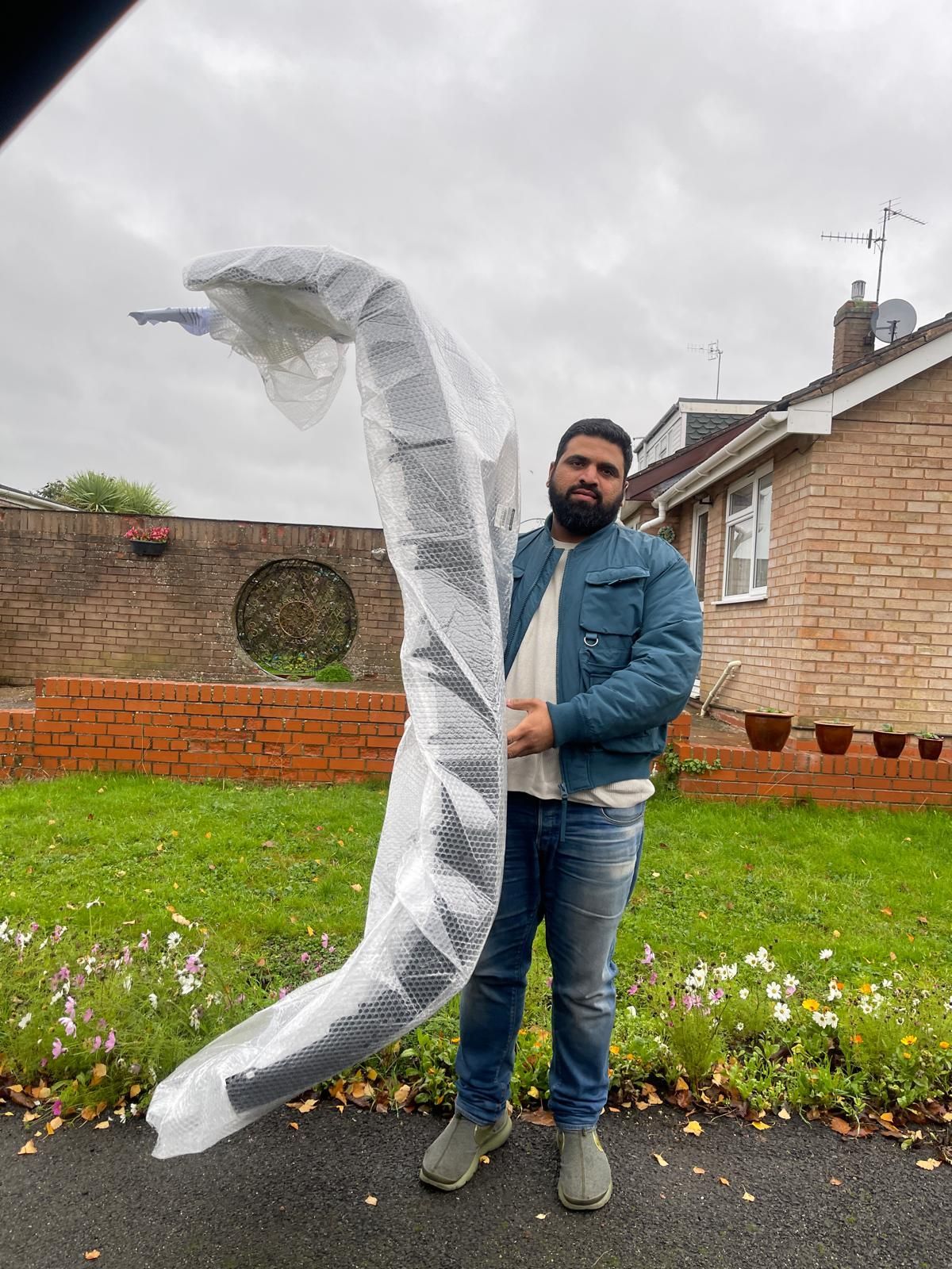 Man holding a long wrapped pole in a garden beside a brick house, under a cloudy sky
