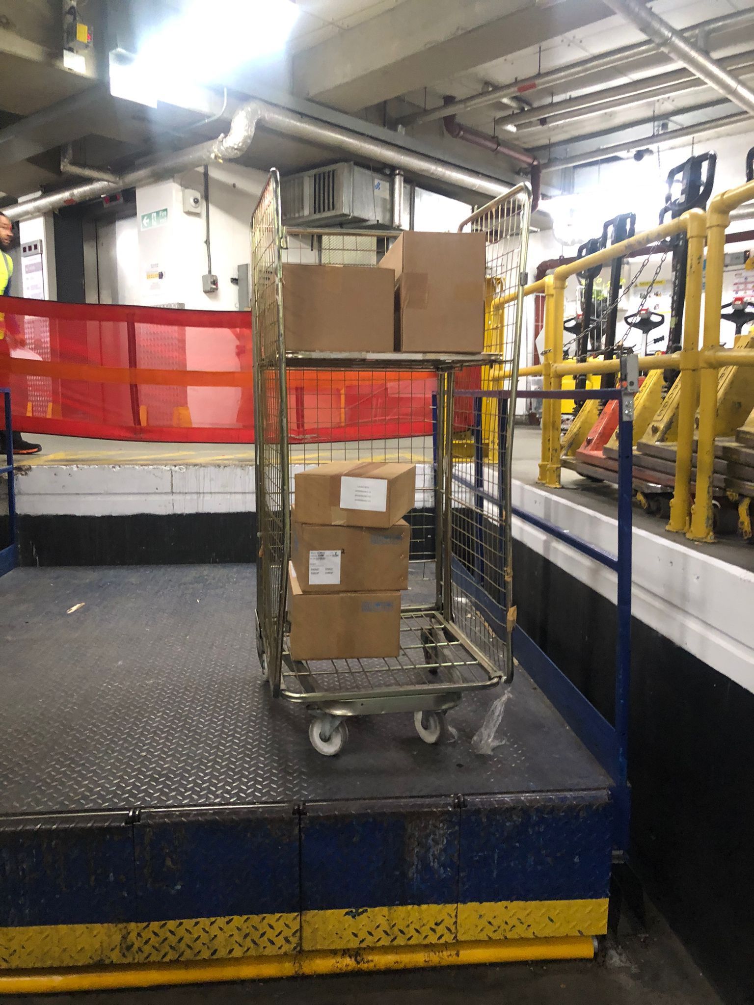 Boxes on a metal cart in an industrial warehouse beside a conveyor and yellow railings.