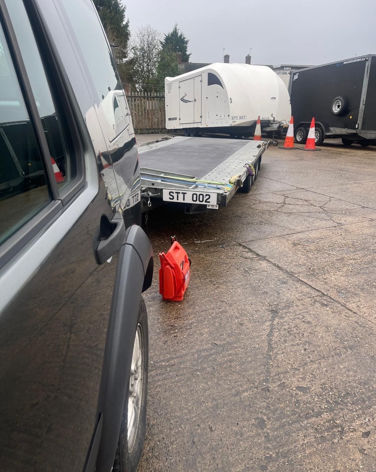 Flatbed trailer beside a van in a gravel lot, with a red gas can in the foreground and cone-marked trailers behind.