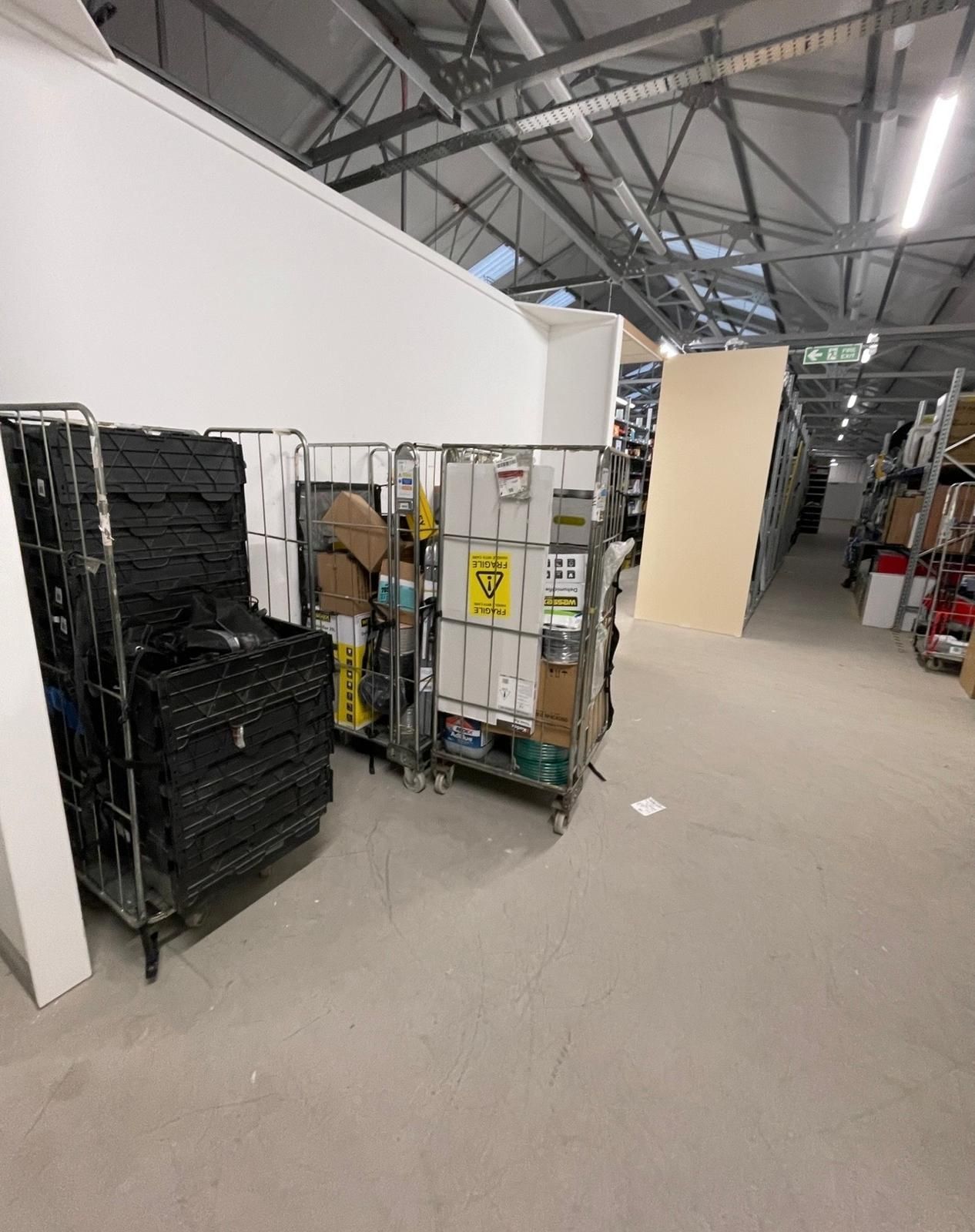 Industrial storage area with stacked black crates, rolling carts, and a white wall under a metal ceiling.