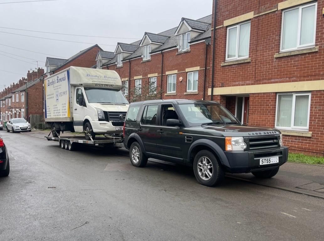 Black SUV towing a white box truck on a wet residential street lined with brick houses.
