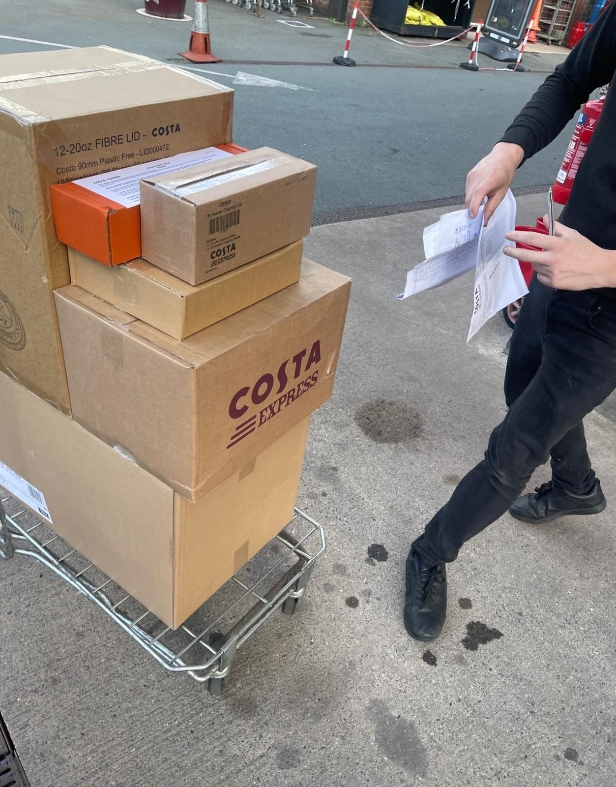 Boxes stacked on a dolly beside a person holding papers on a city sidewalk