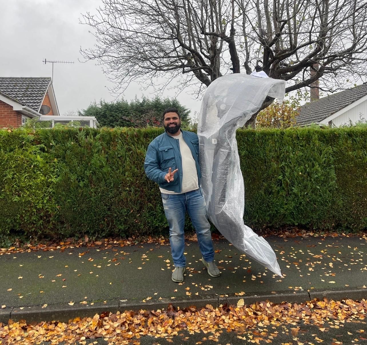 Man posing beside a large transparent inflatable sculpture on a wet sidewalk lined with hedges and autumn leaves