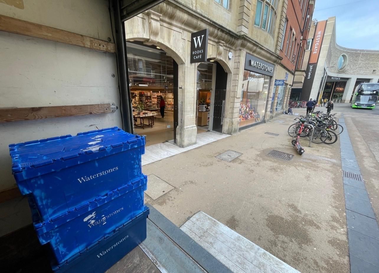 Blue crates outside a shop along a narrow pedestrian street with parked bicycles.
