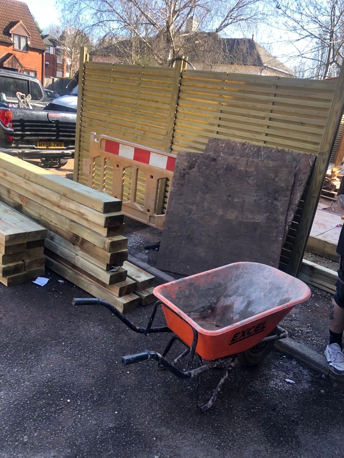 Construction materials stacked by a fence, with a red wheelbarrow and a leaning board on a driveway.