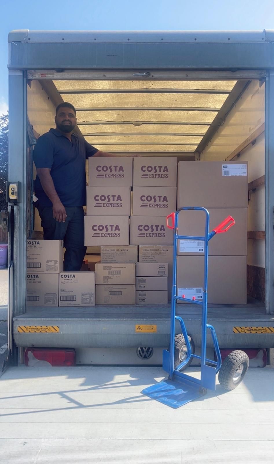 Worker in truck with stacked Costa boxes, unloading alongside a blue hand truck.