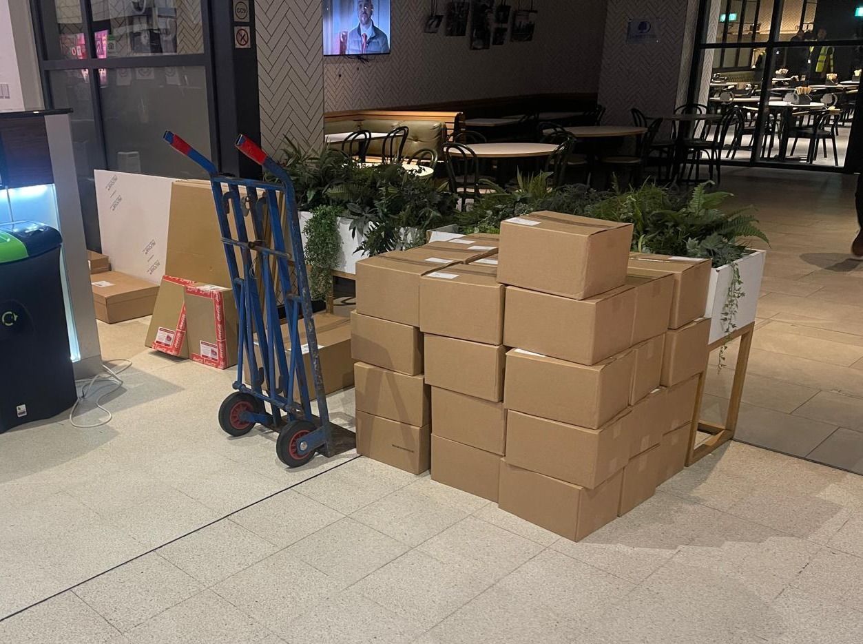 Stacked cardboard boxes beside a hand truck in a building lobby, with plants and a lit storefront in the background