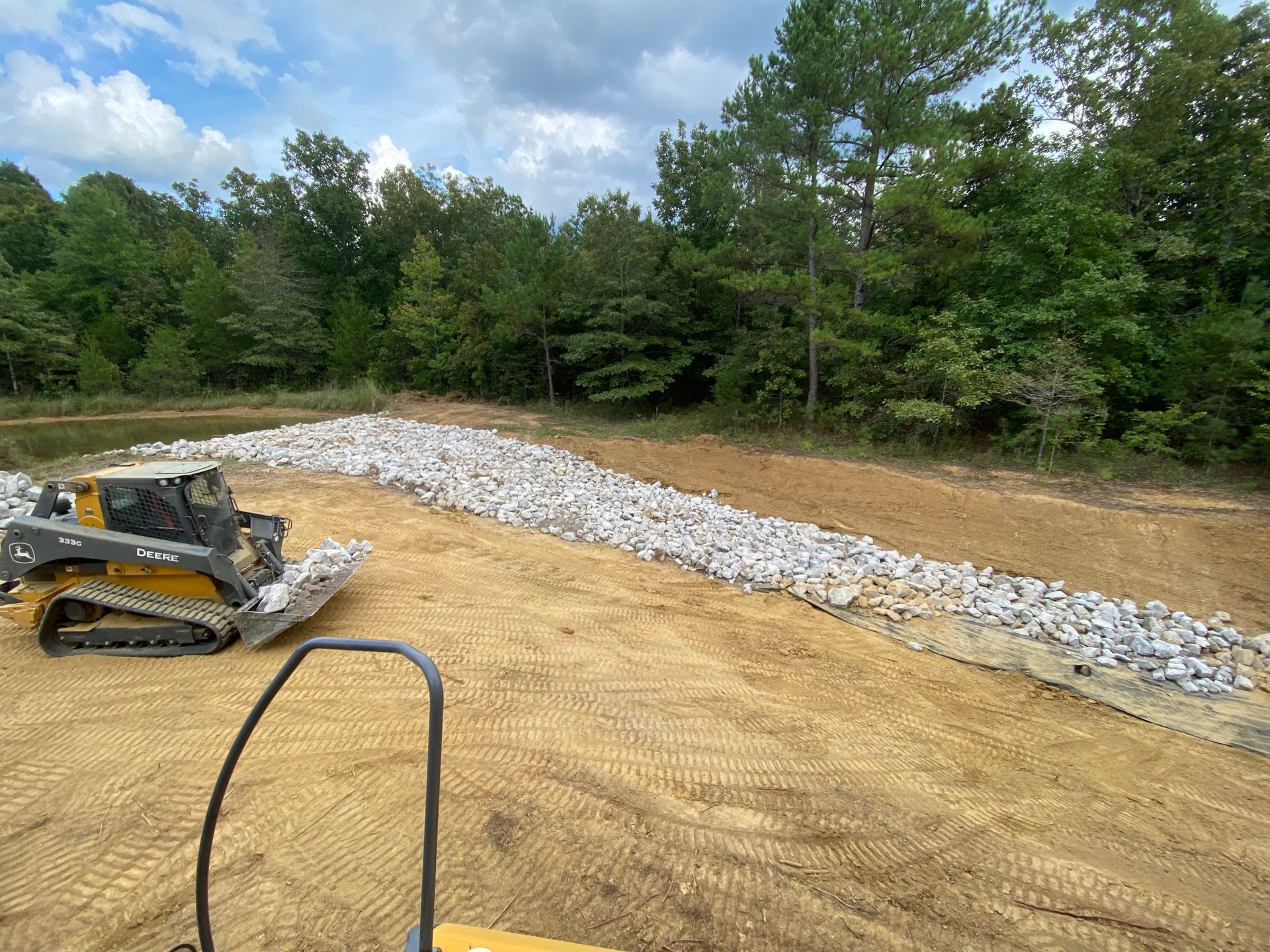 A small construction vehicle laying rocks along a bank of a body of water, near a wooded area.