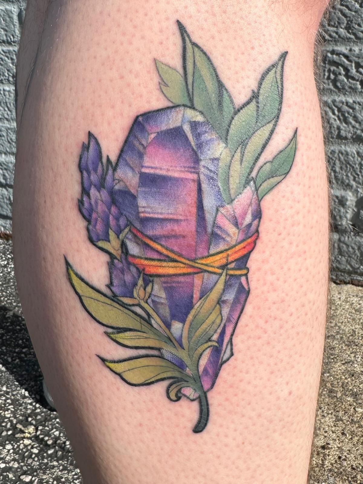 A person has a tattoo of a purple crystal with flowers and leaves on their leg.