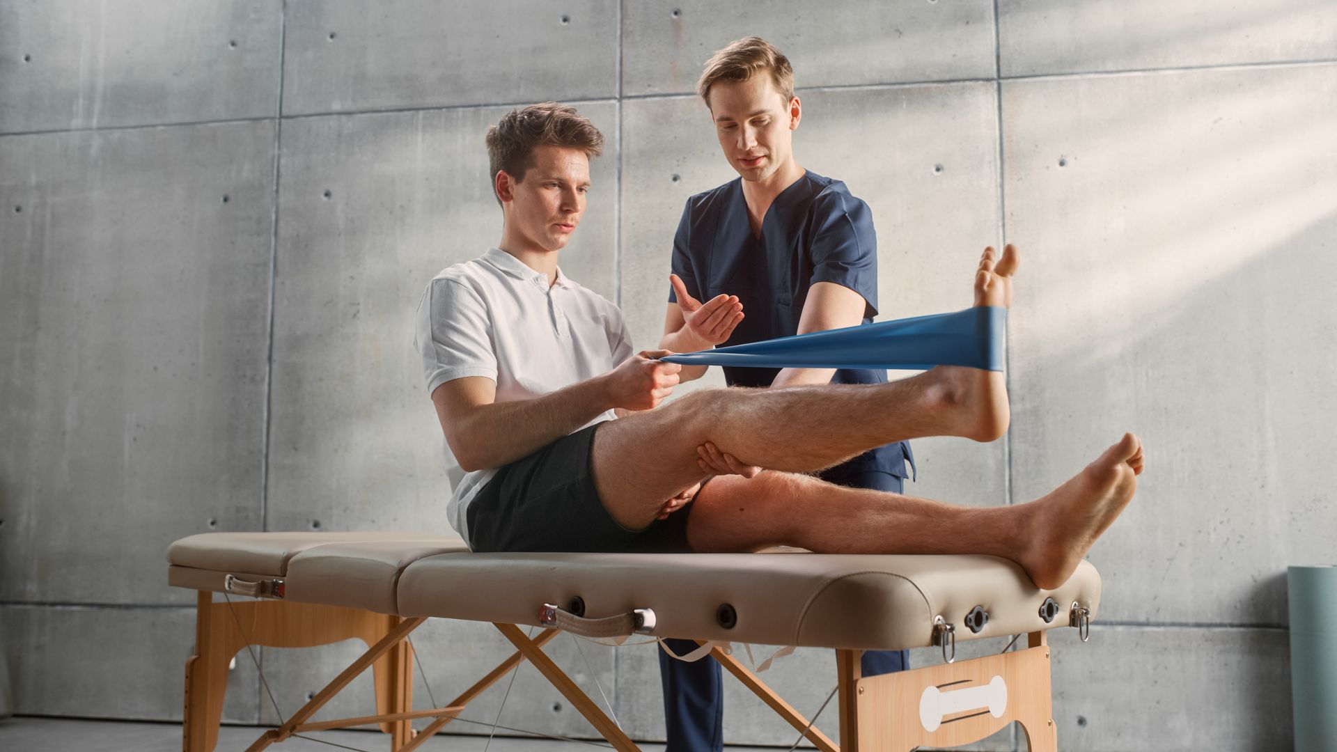 Physical therapist assisting a man doing leg exercises on a treatment table in a bright clinic