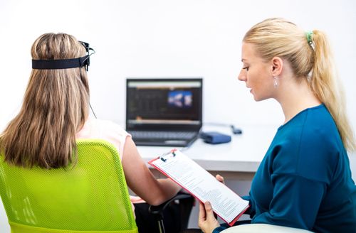 Two people in an office discuss a laptop while one takes notes on a clipboard.