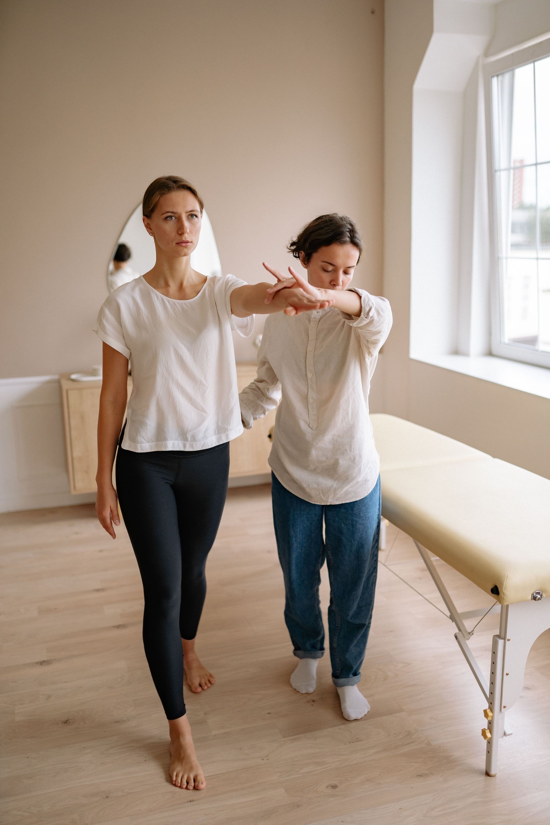 Two people in white shirts walking arm in arm through a bright room with a massage table