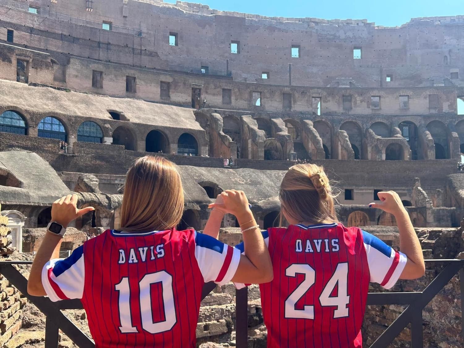 Two fans in red Davis jerseys pose at the Colosseum, raising their arms toward the ancient ruins.