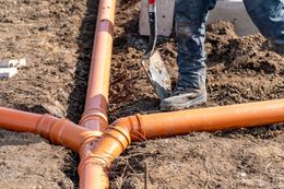 Worker using a shovel to clear dirt around orange PVC drainage pipes laid in a trench.