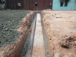 A narrow trench for a concrete foundation, featuring steel rebar reinforcement, sits between leveled gravel and dirt ground.