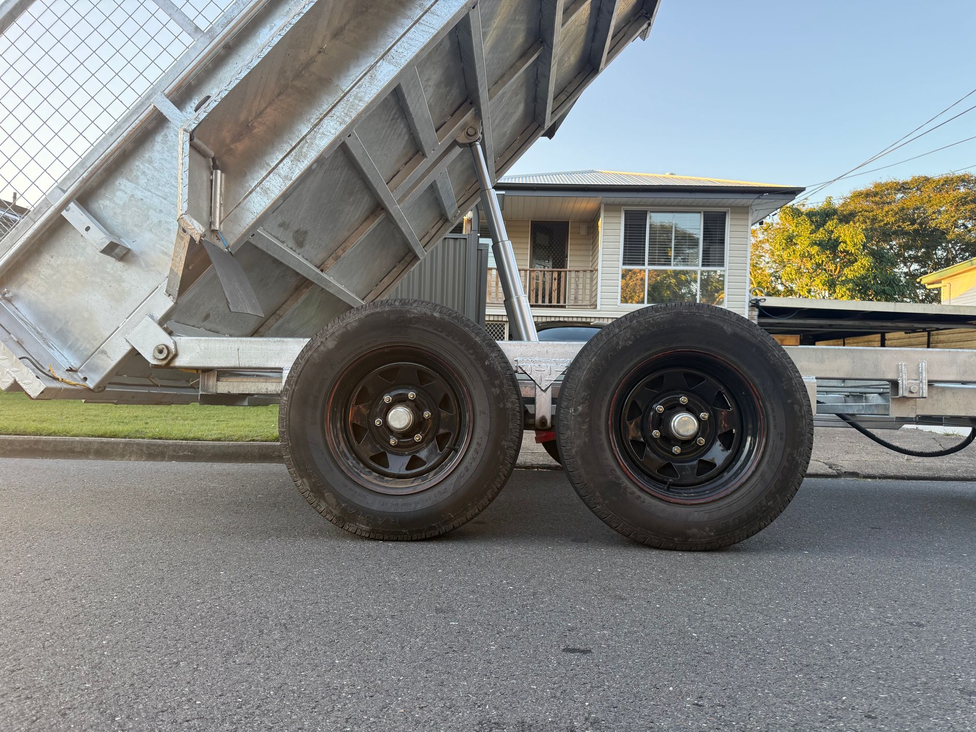 A metal trailer with raised bed, black wheels, parked on asphalt in front of a house.