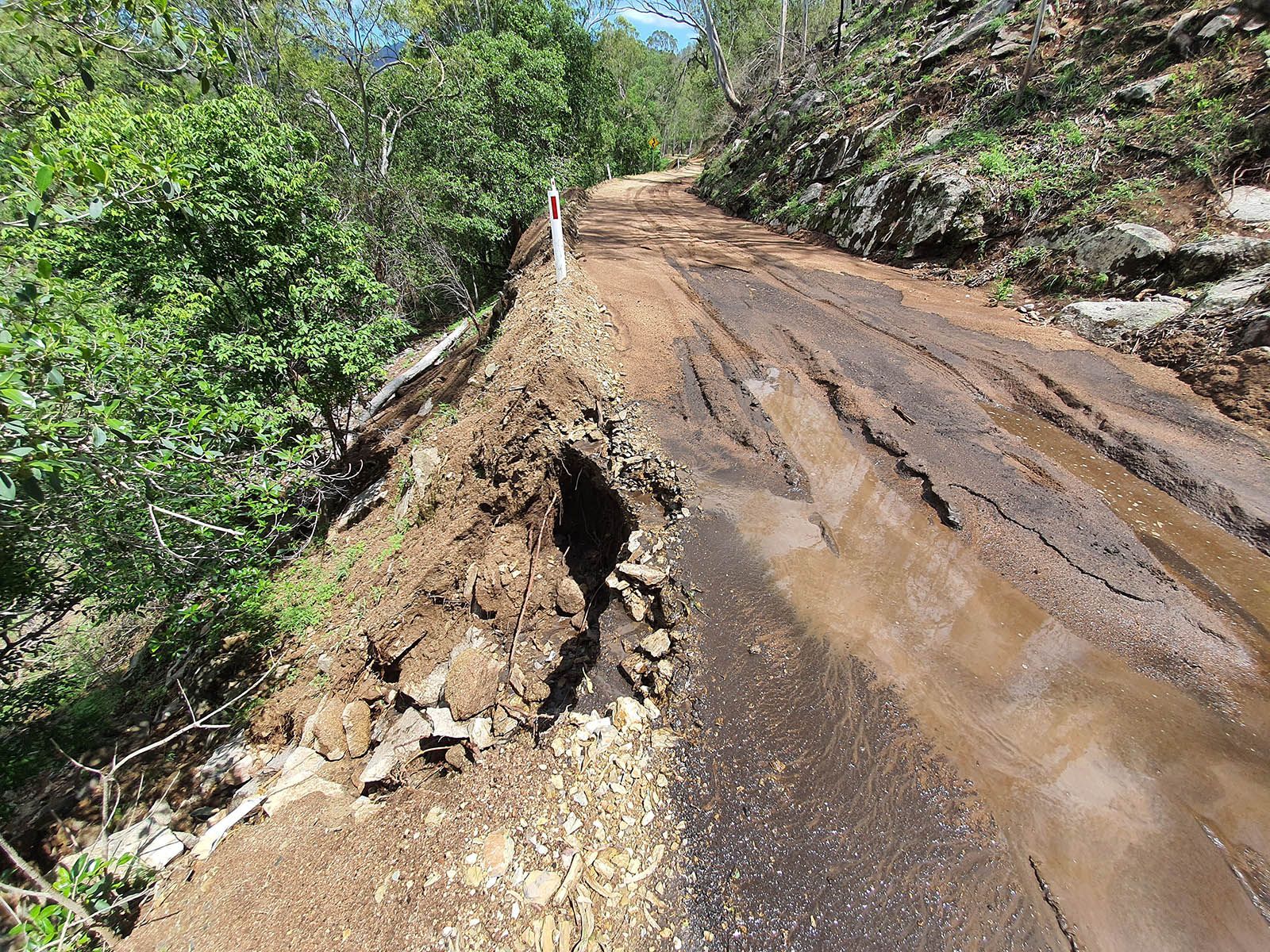 Damaged dirt road washed out by erosion beside a muddy slope and green trees