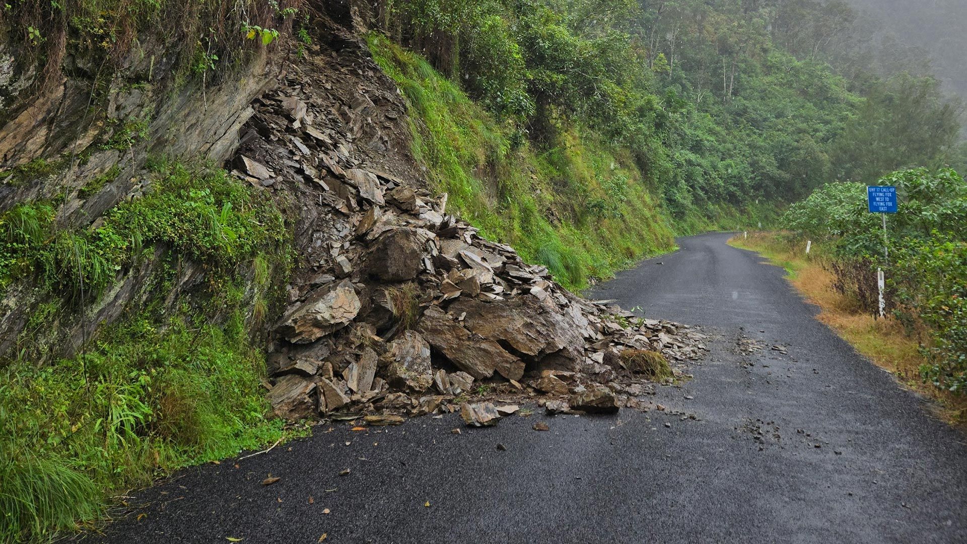 Rockslide blocking part of a wet mountain road beside a grassy hillside
